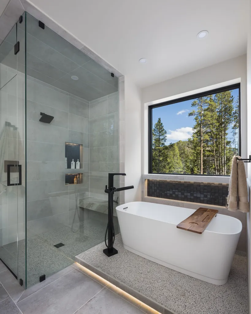 Glass shower and freestanding tub beside a forest-view window