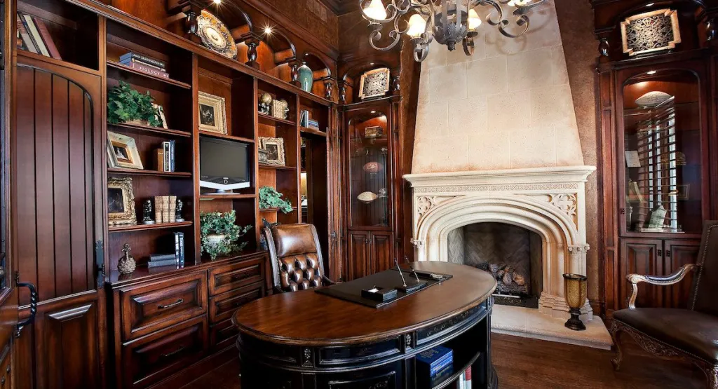 Library with carved wood shelving, a leather-topped desk and a cast-stone fireplace